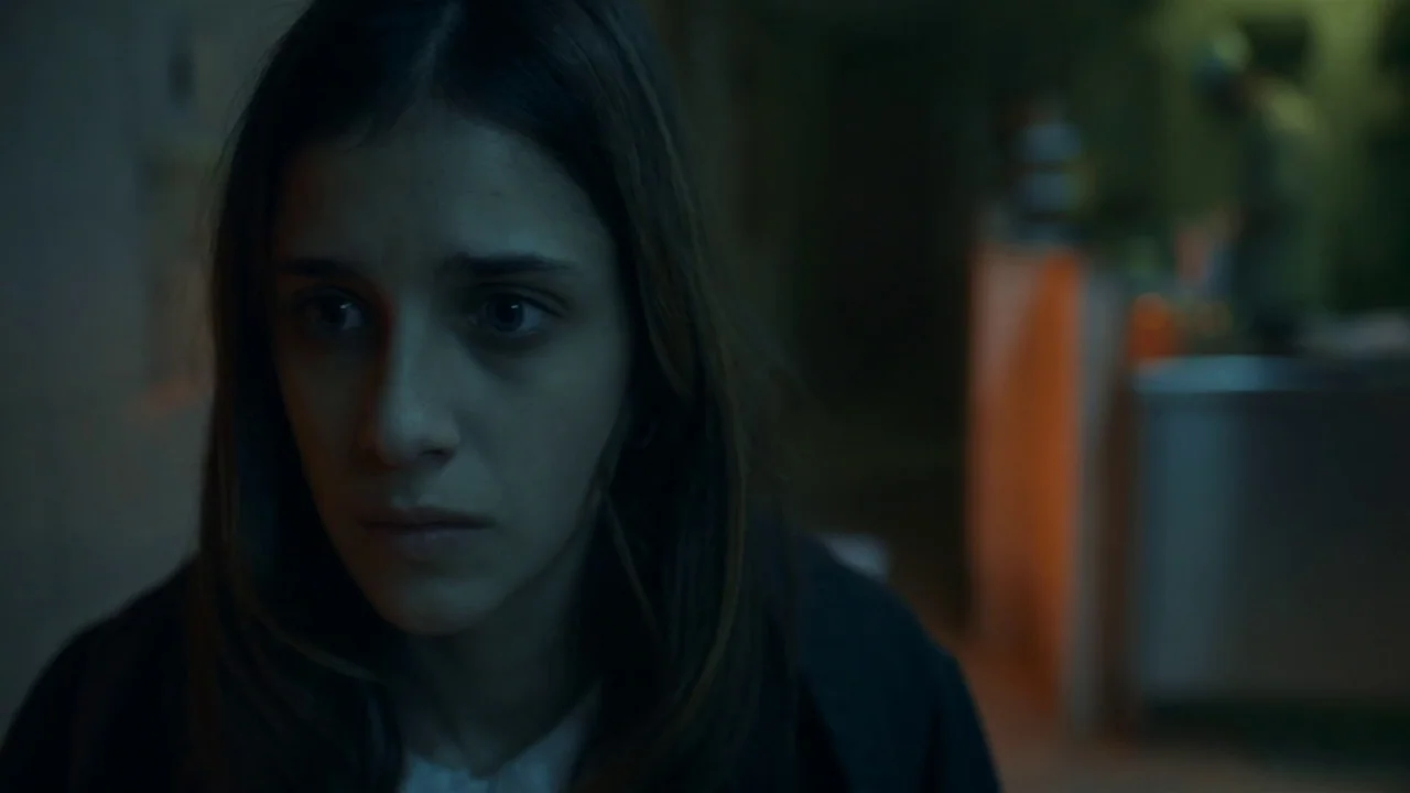 Amanda (2022) movie still: closeup — A dimly lit indoor space features a person in the foreground and blurred machinery. The subject stares into the…
