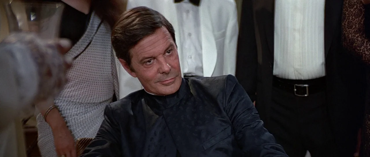 Octopussy (1983) movie still: medium — A formal event interior with attendees wearing black and white evening attire. A man sits at a table and…