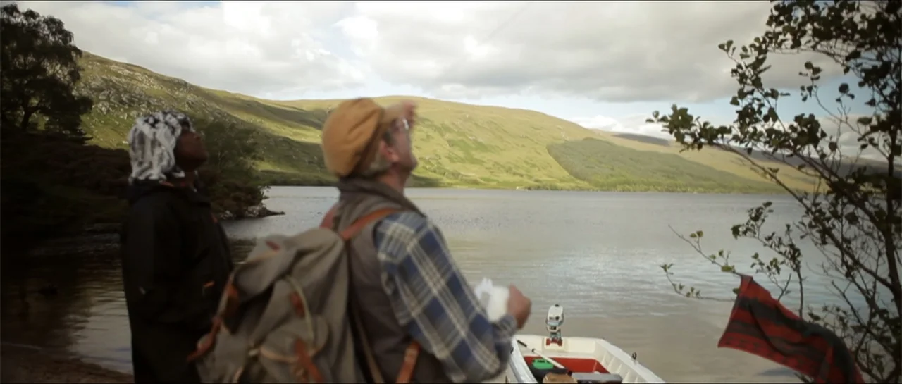 Garnet’s Gold (2014) movie still: medium — Two men stand on the grassy bank of a large lake with green hills beyond. They look up toward the sky as…