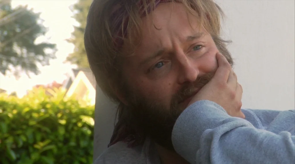 Humpday (2009) movie still: closeup — An outdoor porch area with green hedge foliage and partial house structure in background. A man with a beard…