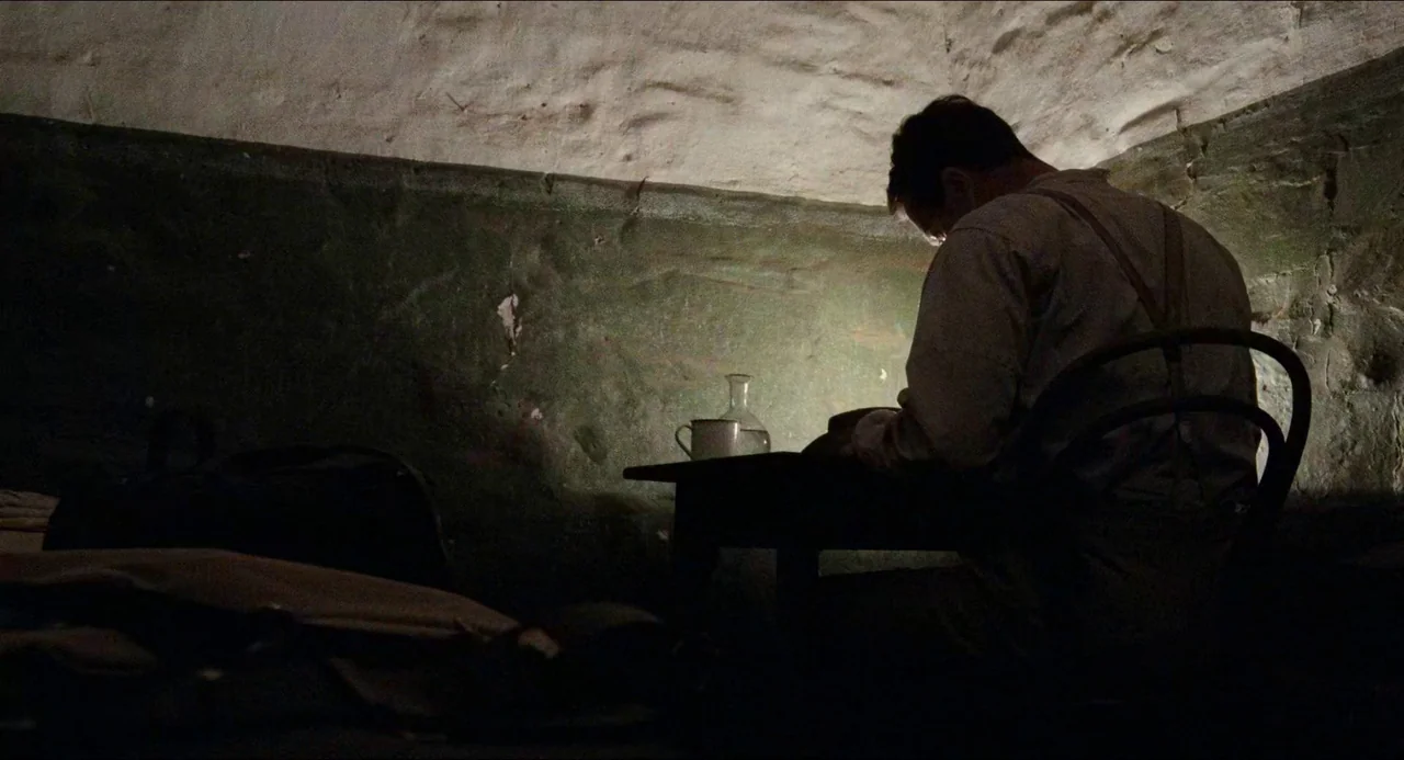 Breaker Morant (1980) movie still: medium — A dark stone room with rough textured walls and minimal furnishings. A man sits at a table writing in a…