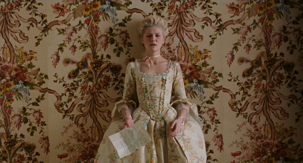 Marie Antoinette (2006) movie still: medium — A woman stands before a wall covered in intricate floral pattern wallpaper. She holds a letter while…
