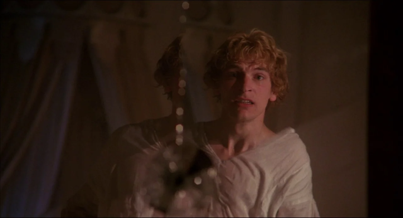 Gothic (1986) movie still: medium — A dimly lit room with draped fabric in the background. A young man with blonde hair gazes forward in a loose…