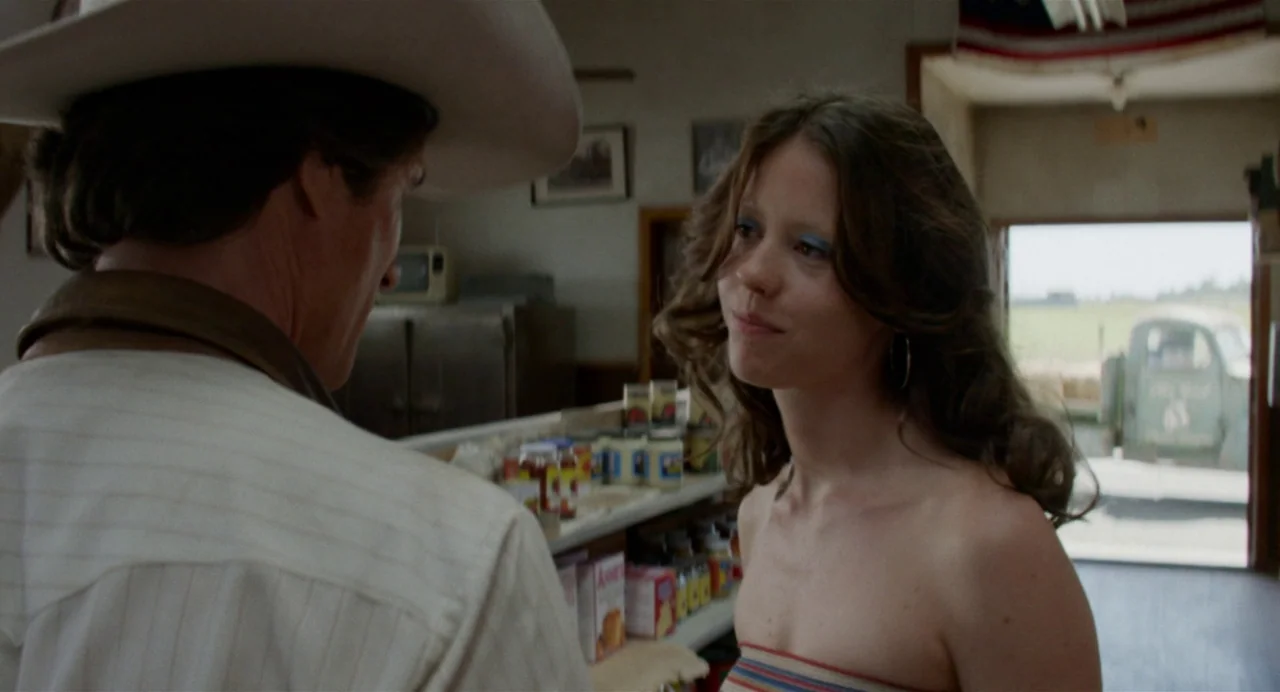 X (2022) movie still: medium — A rustic country store with shelves of canned goods and a wide doorway. A man in a cowboy hat faces a woman with long…