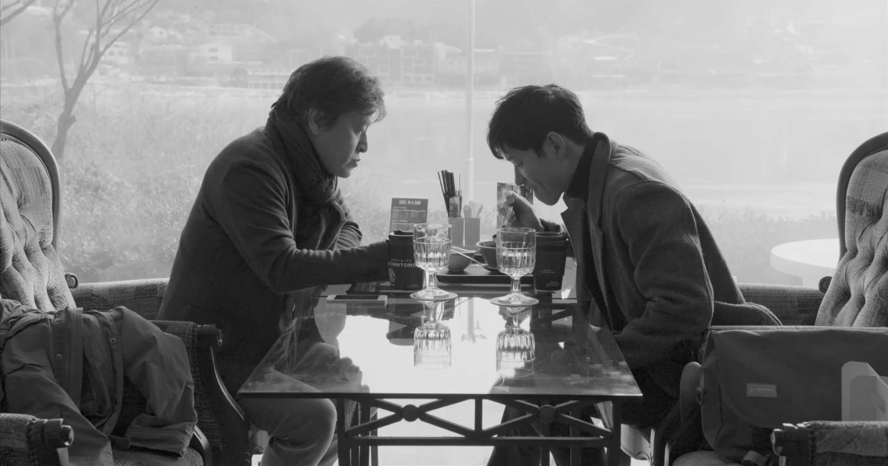 Hotel By The River movie still: medium — Two men sit facing each other at a reflective glass table inside a cafe. Large windows behind them reveal a…