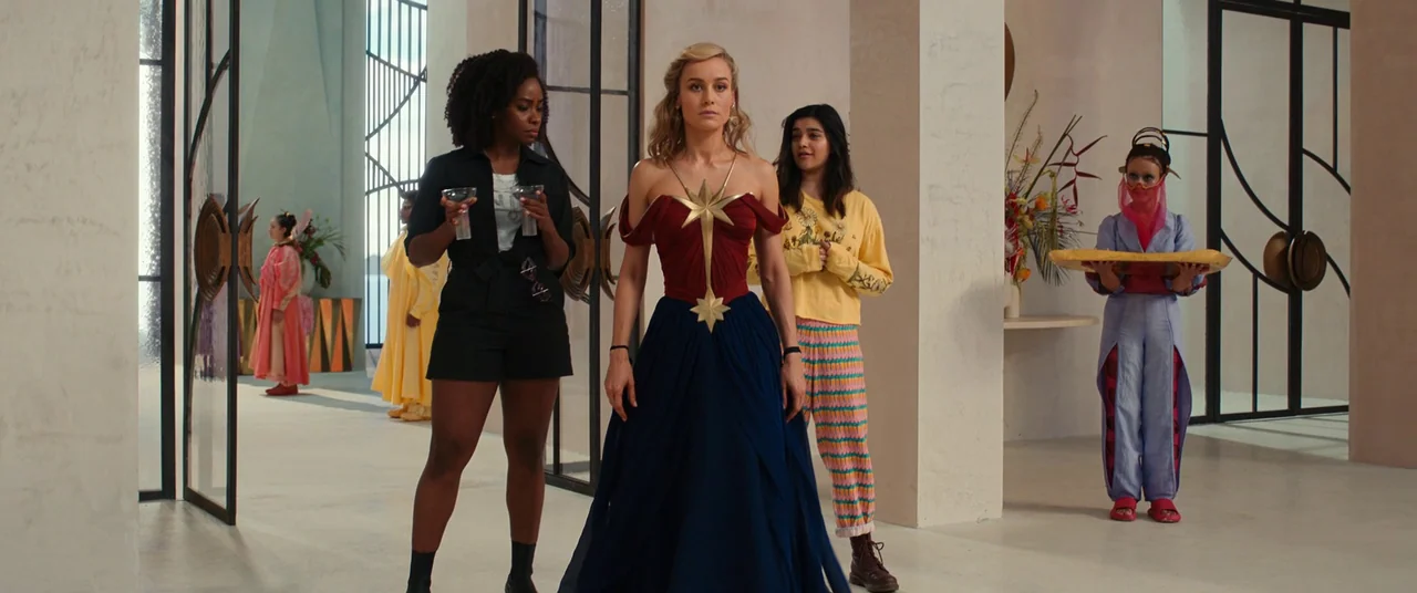 The Marvels (2023) movie still: fullbody — A spacious, minimalist hall with pale walls and floor-to-ceiling glass panel dividers. Five women stand…