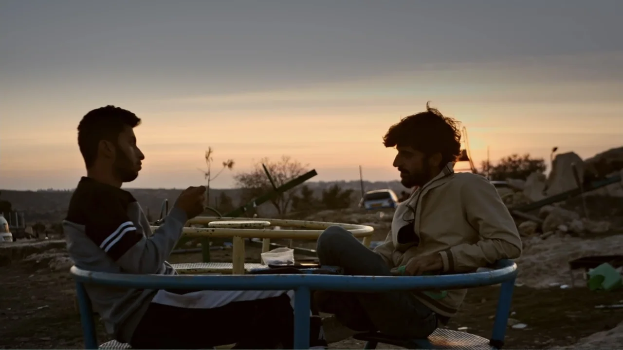 No Other Land (2024) movie still: medium — A playground carousel sits in a desolate exterior area during the golden hour. Two men are seated on the…