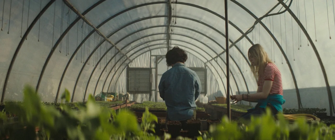 I Think We’re Alone Now (2018) movie still: wide — A plastic-walled agricultural structure filled with rows of starter plant trays. A man and a…