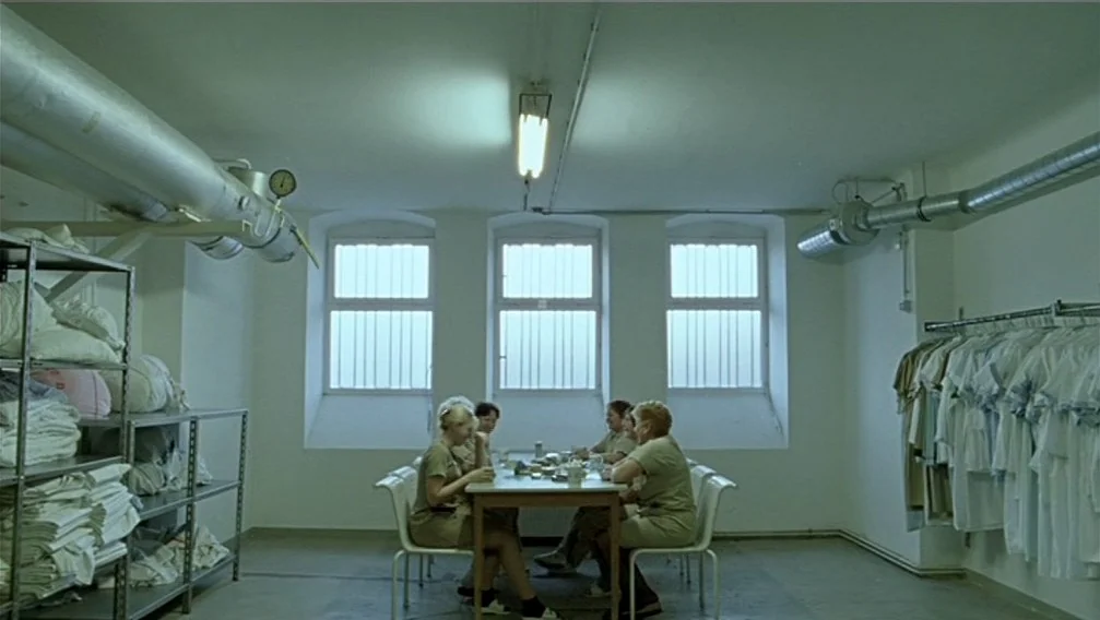 Import Export (2007) movie still: wide — A stark utility room features white walls, metal shelving units, and three windows. Four women in uniform…