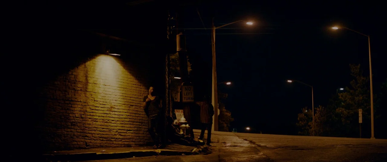 Triple 9 (2016) movie still: wide — A dark urban roadside features a brick wall lit by a single amber lamp. Three men loiter in the shadows near a…