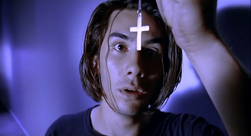 Nowhere (1997) movie still: closeup — A dimly lit interior space with a monochromatic blue color cast. A young man stares directly ahead while…