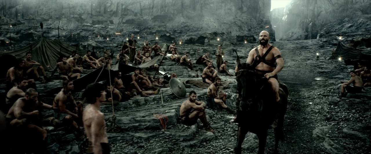 300: Rise of an Empire (2014) movie still: wide — A dark, rocky canyon floor is populated by many shirtless men sitting on stones. A muscular man…