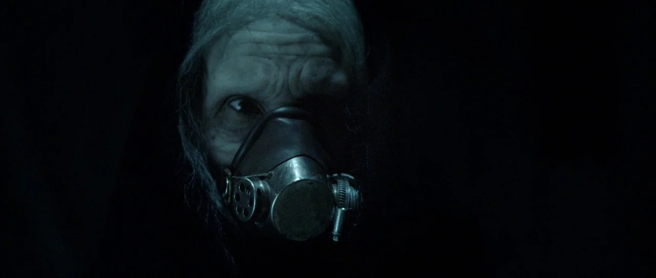 Livid (2011) movie still: closeup — An aged face emerges from deep black shadows with minimal surrounding detail. The subject wears a metallic…