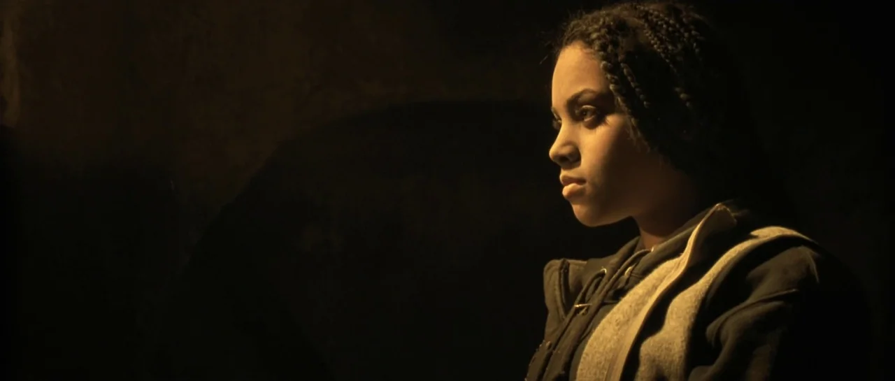 Bones movie still: medium — A dimly lit interior space with a dark background and no visible architectural details. A young woman with braids is…