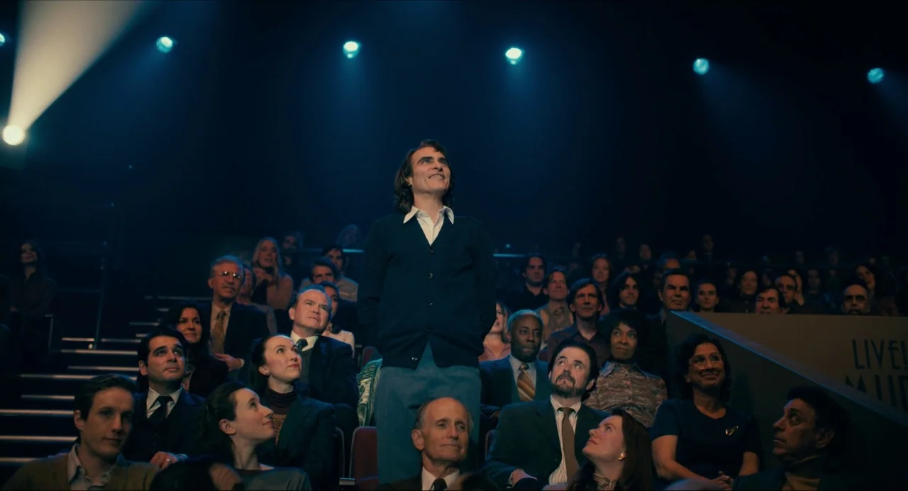 Joker (2019) movie still: medium — A television studio audience sits in tiered dark rows facing the central stage area. A man in a blue cardigan…