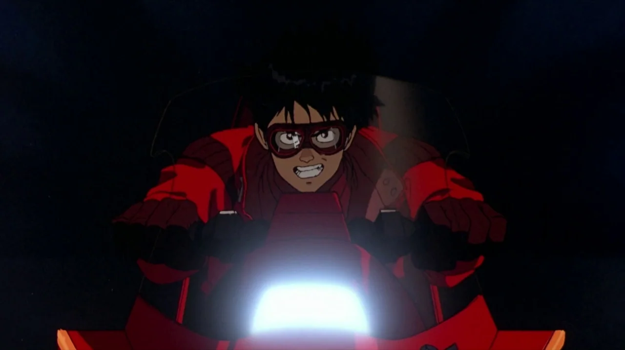 Akira (1988) movie still: medium — A dark night background contrasts with the bright headlight of a high-speed motorcycle. A young man with black…