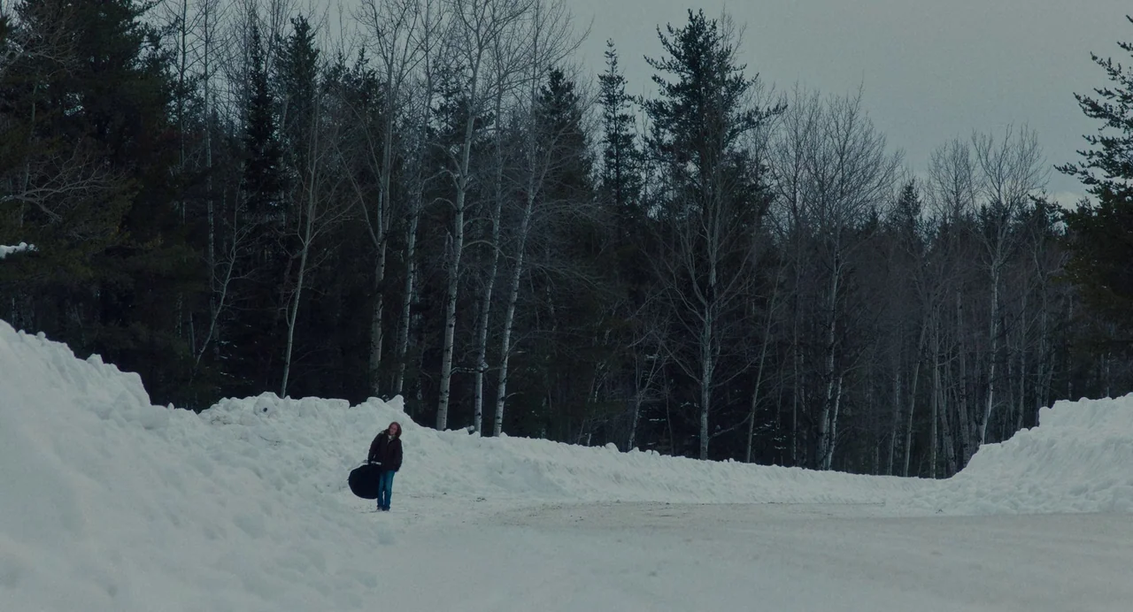 My Animal (2023) movie still: wide — A snow-covered road is flanked by high snow banks and dense bare birch trees. A lone woman walks along the side…