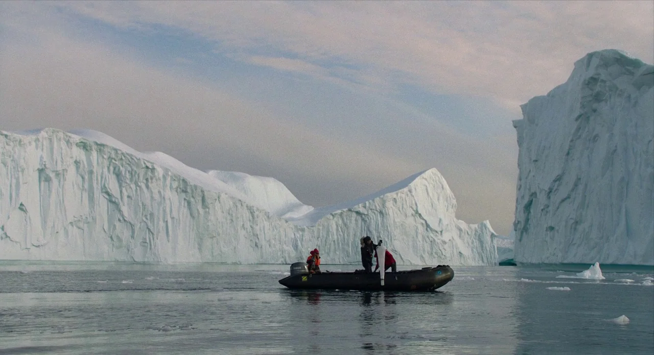 Where’d You Go Bernadette (2019) movie still: wide — Massive glacial ice shelves loom over a small dark inflatable motorboat in arctic water. The…