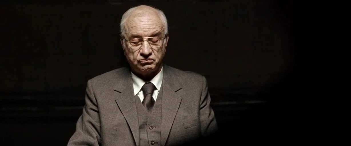 The International (2009) movie still: medium — A man sits against a completely dark, featureless background that suggests an isolated interior…