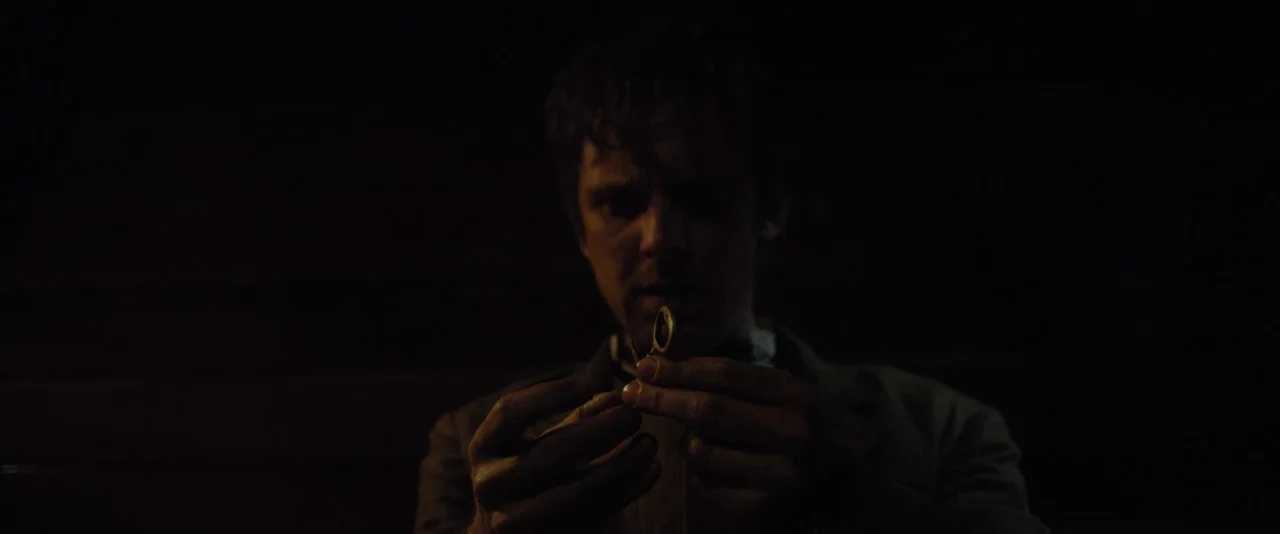 Apostle (2018) movie still: medium — A dimly lit interior space with almost entirely obscured background details and deep shadows. A man in a suit…