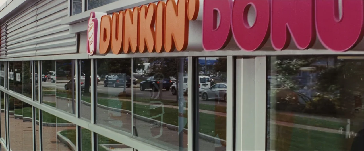 Unfinished Business (2015) movie still: medium — A modern commercial building exterior features a bright orange and pink brand sign. Glass panels…