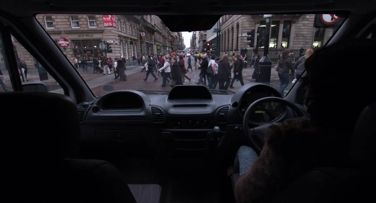 Under The Skin (2013) movie still: wide — The interior of a vehicle dashboard overlooks a crowded city intersection with pedestrians crossing. Many…