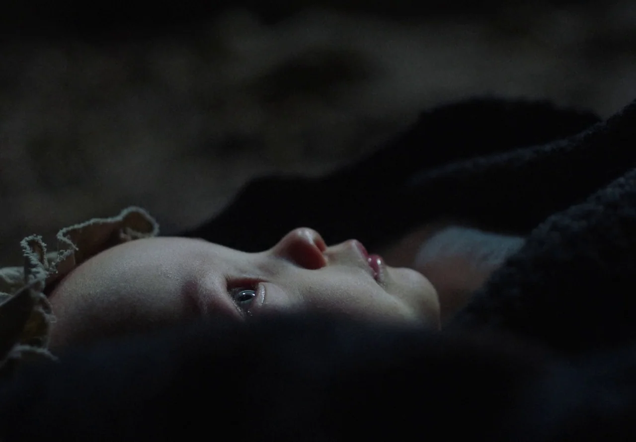 You Won’t Be Alone (2022) movie still: closeup — An infant rests on a dark surface, wearing a simple fabric bonnet. The face is illuminated by a…