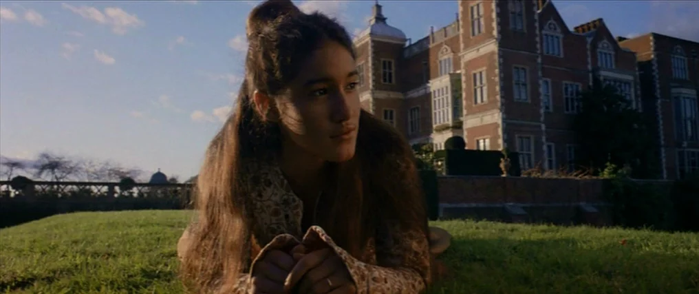 The New World (2005) movie still: medium — A sprawling brick mansion stands behind a neatly mowed grassy lawn at dusk. A young woman rests in the…