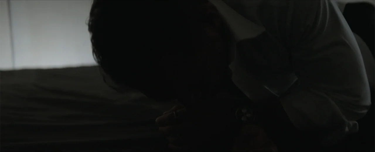 Venom: The Last Dance (2024) movie still: closeup — A dark, minimally furnished interior space features a person in a white shirt. The subject leans…