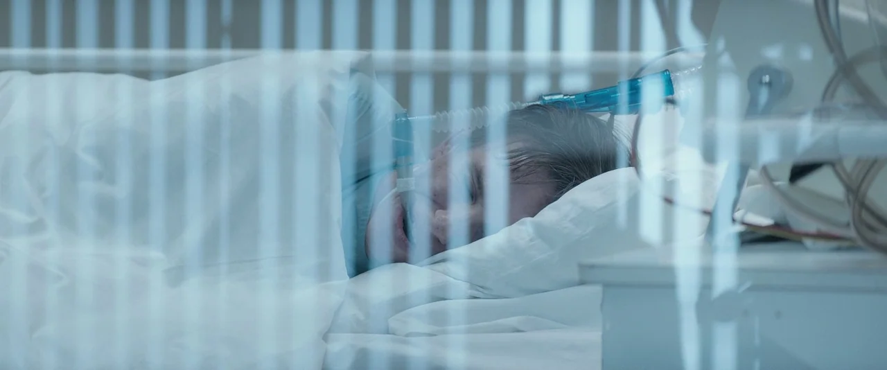 The Theory of Everything (2014) movie still: closeup — A hospital patient lies in a bed behind a glass partition with vertical blinds. The patient…