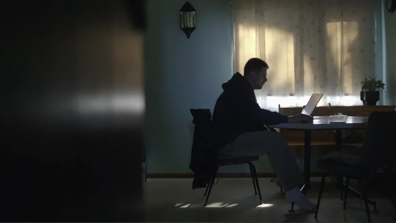 Beware the Slenderman (2016) movie still: medium — A residential room features a circular table situated before a bright, draped window. A man sits…