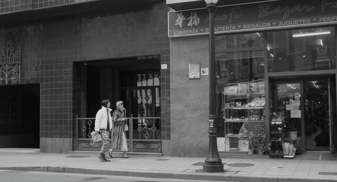 El Planeta (2021) movie still: fullbody — A pedestrian sidewalk passes by a storefront with a large glass window display. Two women walk together…