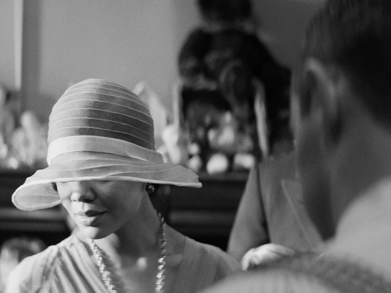 Passing (2021) movie still: closeup — An interior space with a blurred figure in the background. A woman wearing a 1920s style cloche hat looks…