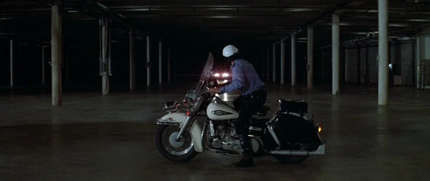 Brewster McCloud (1970) movie still: wide — A vast, empty concrete parking structure features repetitive white structural columns in low light. A…