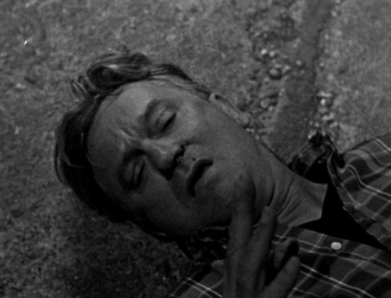 Bait (2019) movie still: closeup — A man lies on coarse asphalt. His eyes are shut and he is motionless.