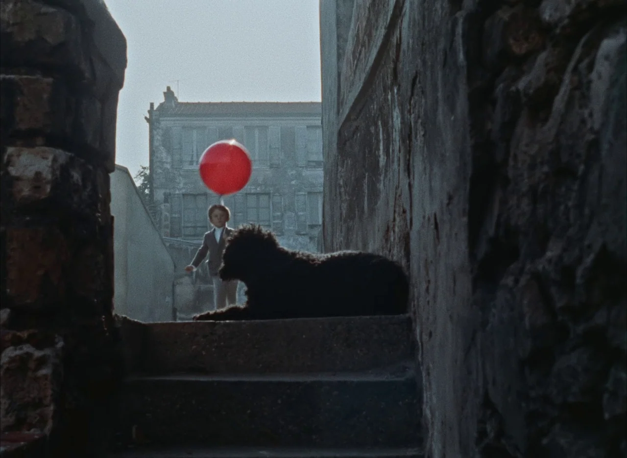 The Red Balloon (1956) movie still: wide — A narrow cobblestone alleyway sits between weathered stone walls. A small boy walks with a red balloon…