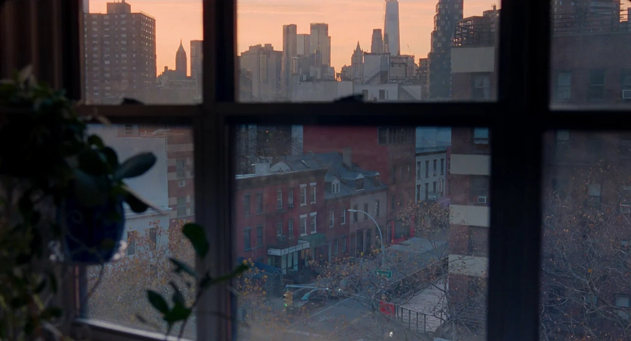 His Three Daughters (2023) movie still: establishing — An interior view through a window shows a densely packed city street below. Tall skyscrapers…