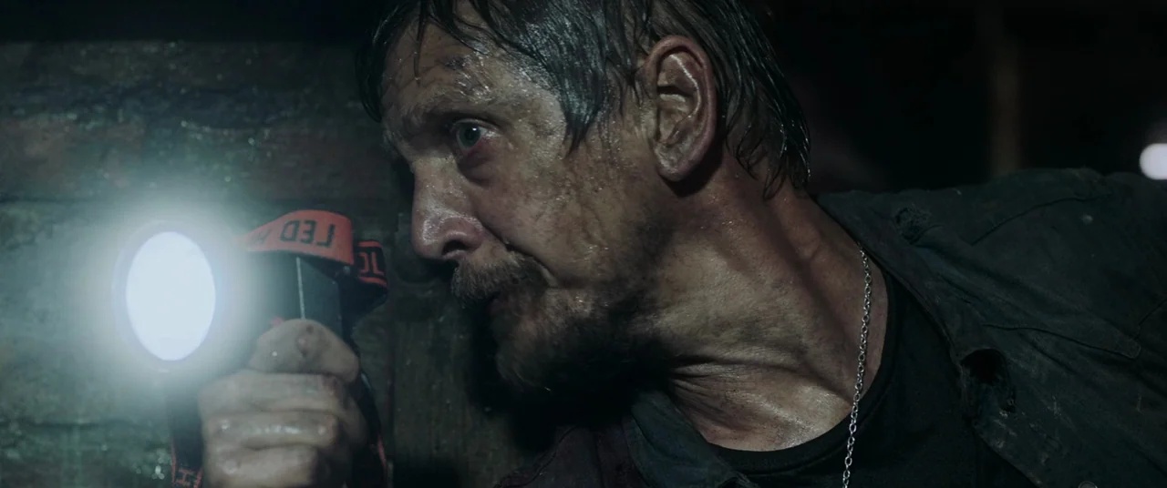 Crawl (2019) movie still: closeup — A dark, cramped stone tunnel with moisture on the walls. A man holds a bright headlamp forward to see in the dark.
