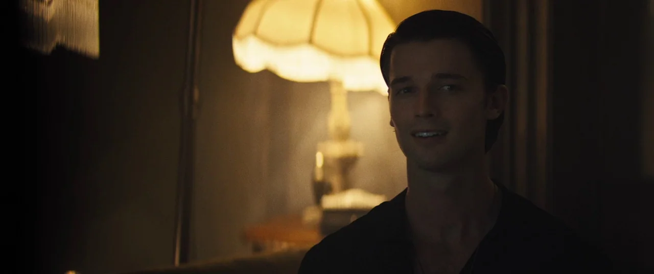 Daniel Isn’t Real movie still: medium — A dimly lit domestic interior features a lit table lamp in the background. A man stands in the foreground,…