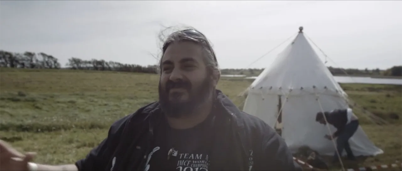 Bludgeon (2018) movie still: medium — A grassy open field contains a white conical tent in the background. A man in a dark jacket stands in the…