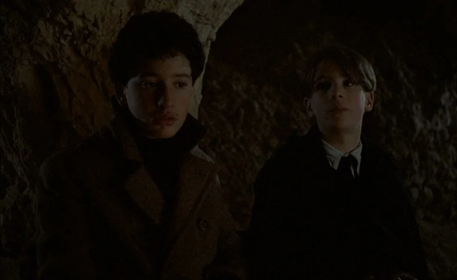 Au Revoir Les Enfants (1987) movie still: medium — Two boys are positioned within a dark, rough-hewn limestone cave interior. The subjects sit near…