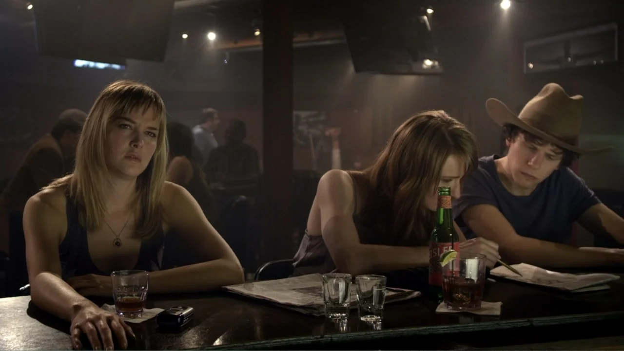 Free Samples (2013) movie still: medium — A dark, wood-paneled bar interior features patrons seated along the counter at night. Two women and one…