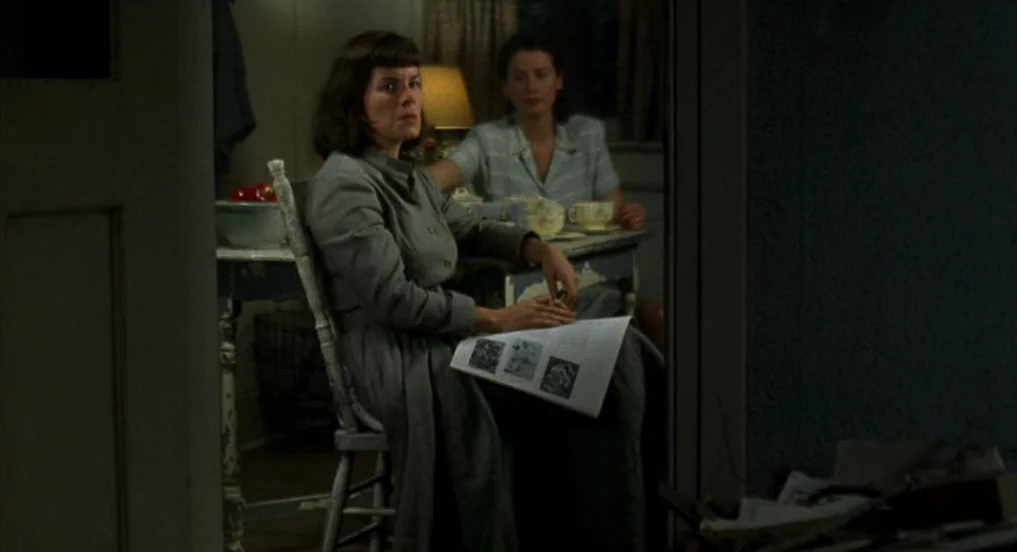 Pollock (2000) movie still: medium — A dimly lit room with a wooden table where two women sit together. One woman sits in the foreground holding…