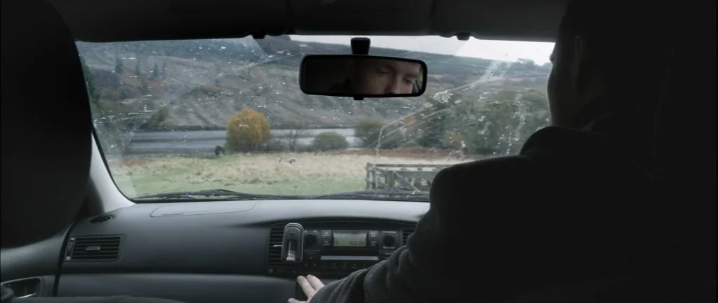 One Hundred Mornings (2009) movie still: medium — A view from the back seat of a modern car looking through the windshield. The driver reaches…