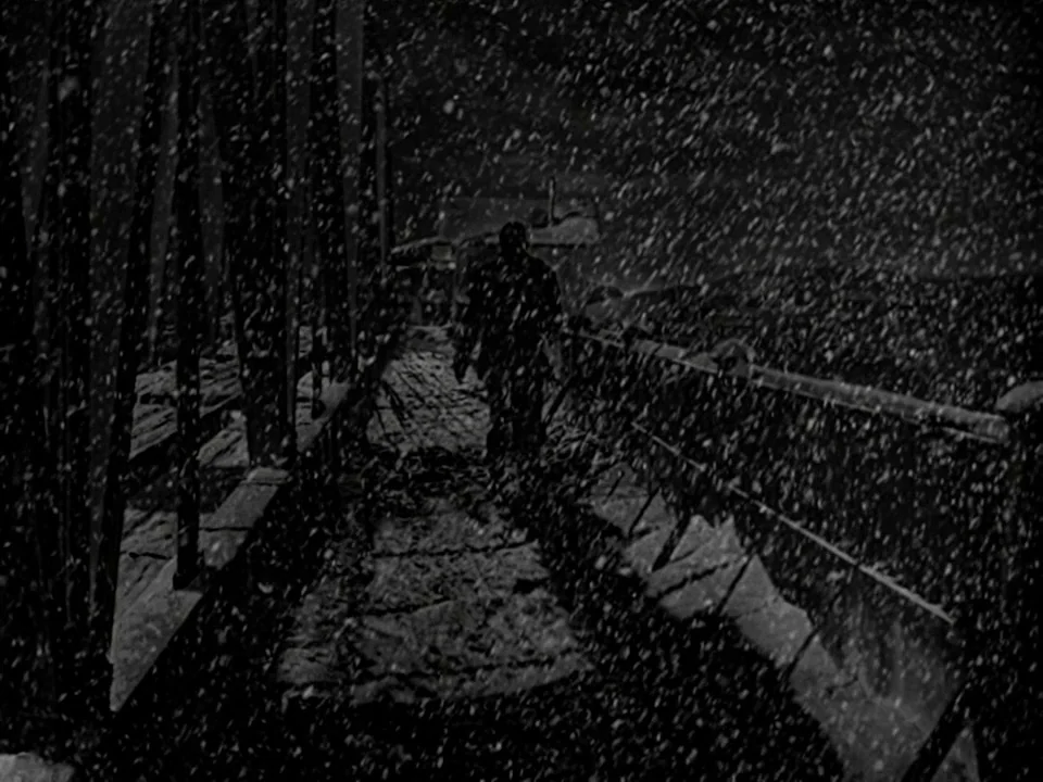 It’s A Wonderful Life (1946) movie still: wide — An industrial metal bridge at night during a heavy blizzard. A lone figure walks away from the…