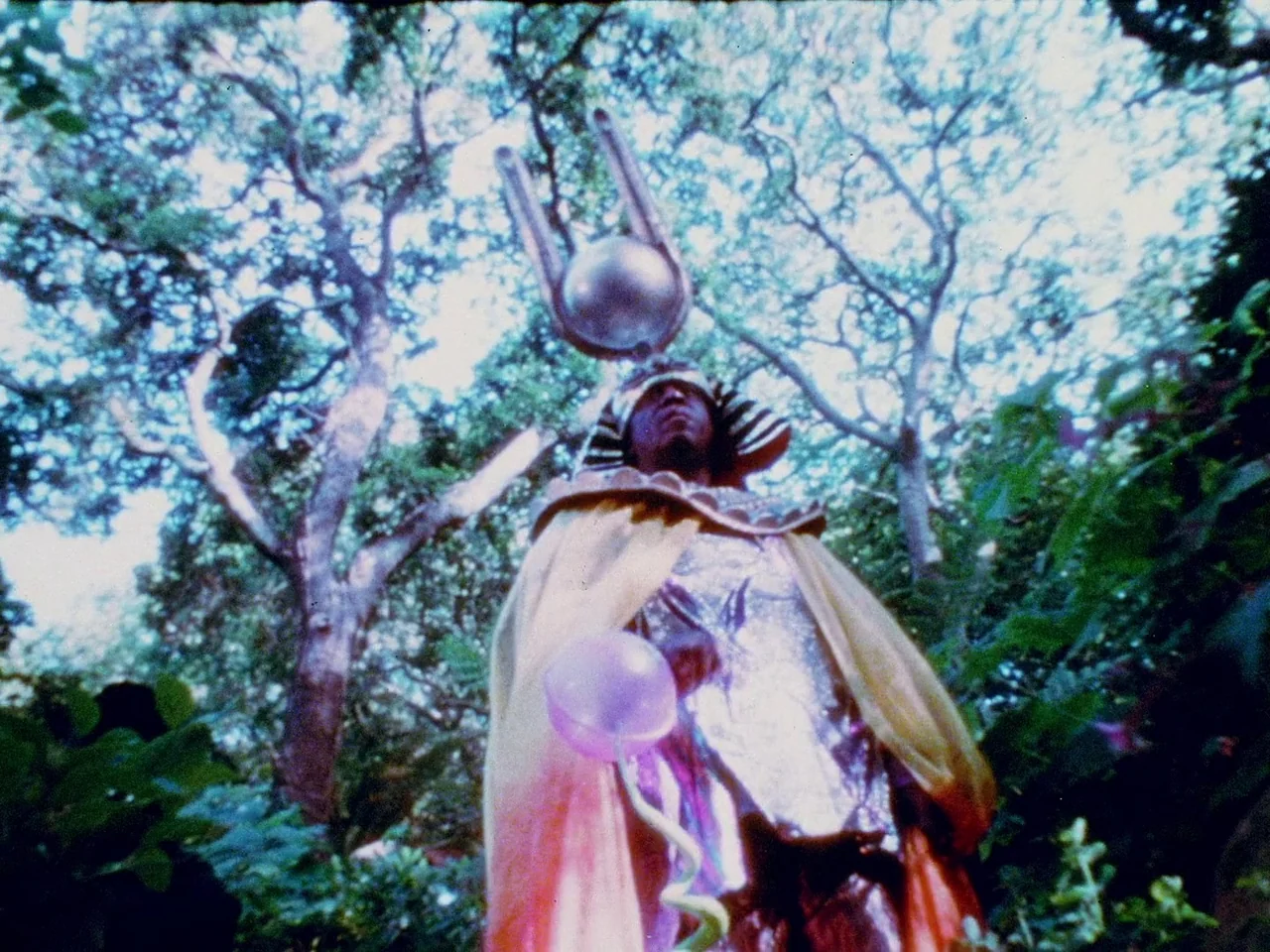 Space is the Place (1974) movie still: fullbody — A person stands in a leafy woodland area beneath tall, sprawling deciduous trees. They wear an…