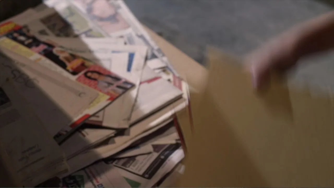 Jinxed (2013) movie still: closeup — A pile of assorted papers and magazines rests on a flat wooden surface. A blurry hand holds a manila envelope…