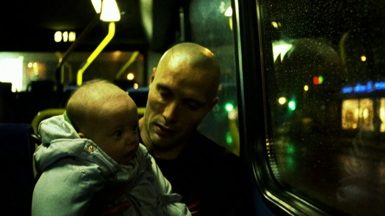 Pusher II (2004) movie still: medium — Interior of a bus at night featuring rain-streaked windows showing blurry city street lights. A man with a…