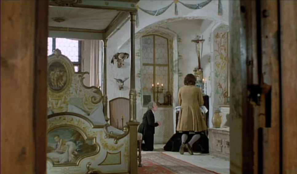 Heart of Glass (1976) movie still: wide — An ornate room in a castle features a large painted bed and stone arches. Two men are kneeling on the…