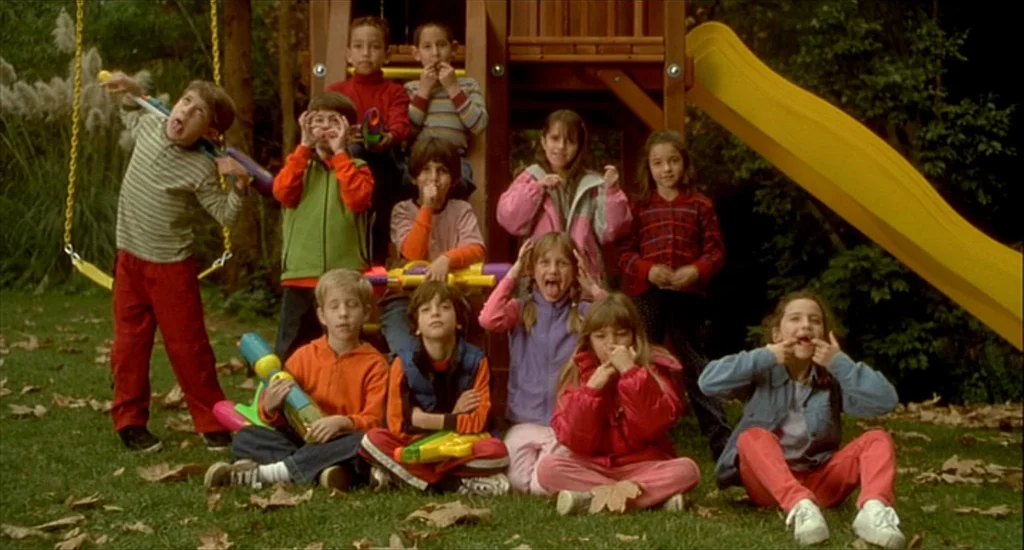 One Hour Photo (2002) movie still: fullbody — A group of children gather around a wooden playground set in a leaf-covered backyard. The children…